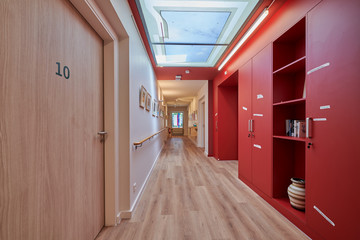 Ein heller Flur mit Holzboden, roten Wandelementen und Schränken und Tageslicht durch Lichtkuppeln im Stationären Diakonie Hospiz Wannsee in Berlin-Wannsee. Ein heller Flur mit Holzboden, roten Wandelementen und Schränken und Tageslicht durch Lichtkuppeln im Stationären Diakonie Hospiz Wannsee in Berlin-Wannsee.