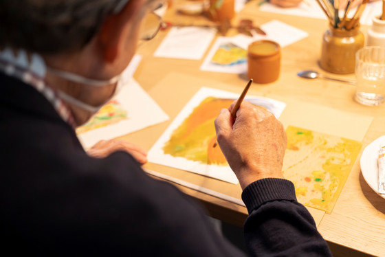 Ein Hospizgast sitzt an einem Tisch in einem mit warmen Licht beleuchteten Aufenthaltsraum mit Malpinseln, Stiften und anderen Malutensilien und zeichnet mit einem Pinsel ein Bild bei der Kunsttherapie im Diakonie Hospiz Wannsee in Berlin-Wannsee. Ein Hospizgast sitzt an einem Tisch in einem mit warmen Licht beleuchteten Aufenthaltsraum mit Malpinseln, Stiften und anderen Malutensilien und zeichnet mit einem Pinsel ein Bild bei der Kunsttherapie im Diakonie Hospiz Wannsee in Berlin-Wannsee.