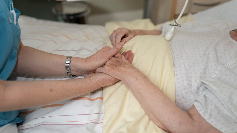 Pflegekraft berührt Hände eines Gastes, der im Bett liegt - Würdiges Sterben - Diakonie Hospiz Wannsee