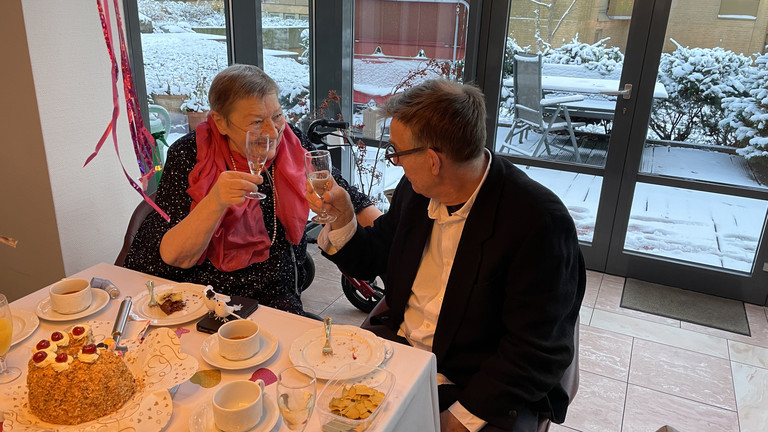 Das Hochzeitspaar sitzt am Kaffee-Tisch und stößt mit einem Glas Sekt an, besonderer Momente im Hospiz, Lebensabschnitt feiern, Diakonie Hospiz Wannsee, Berlin-Wannsee
