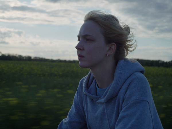 Filmausschnitt aus dem Kinofilm "Sechswochenamt" mit einer Frau vor einem mit Gras bewachsenen Hügel und einem Himmel mit Wolken.