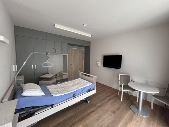 Blick in ein helles Gästezimmer mit einem frisch bezogenen Gästebett, Holzboden, hellen Wandfarben und Monitor an der Wand im Stationären Diakonie Hospiz Wannsee in Berlin-Wannsee.